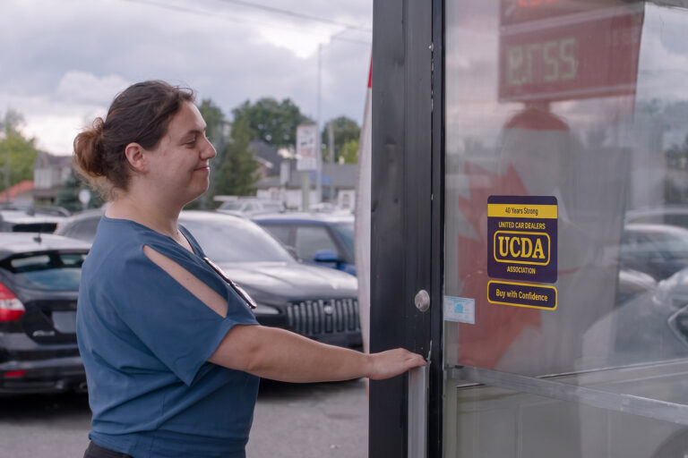 A young woman walks up to a used car dealership’s door showing the blue UCDA sign